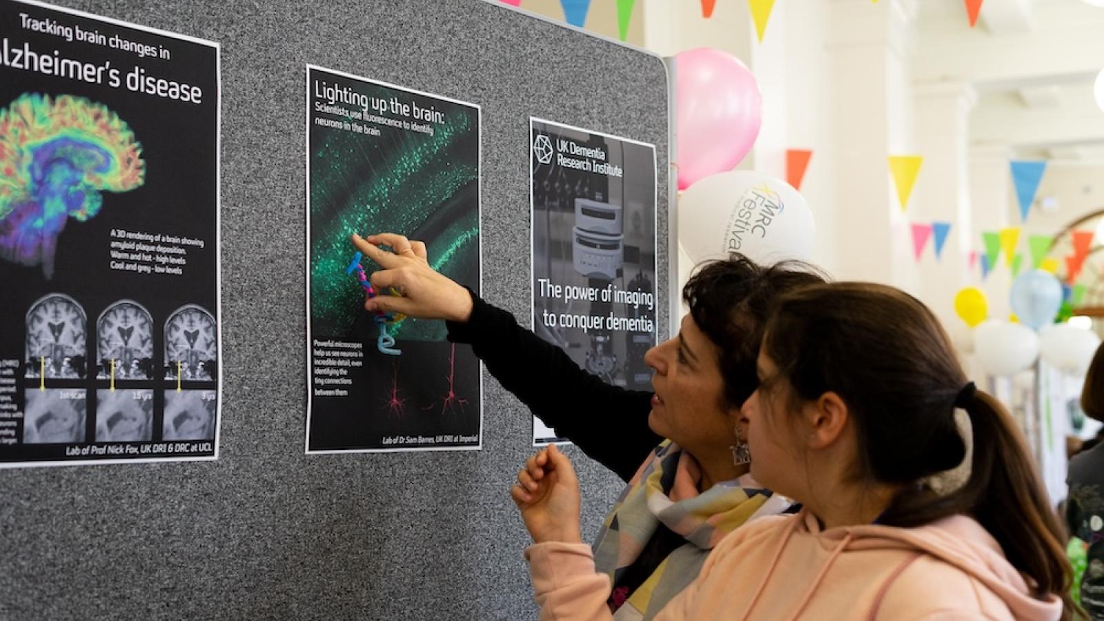 Two women examine research posters about Alzheimer's disease and brain imaging at a festive event with colourful balloons and bunting in the background.