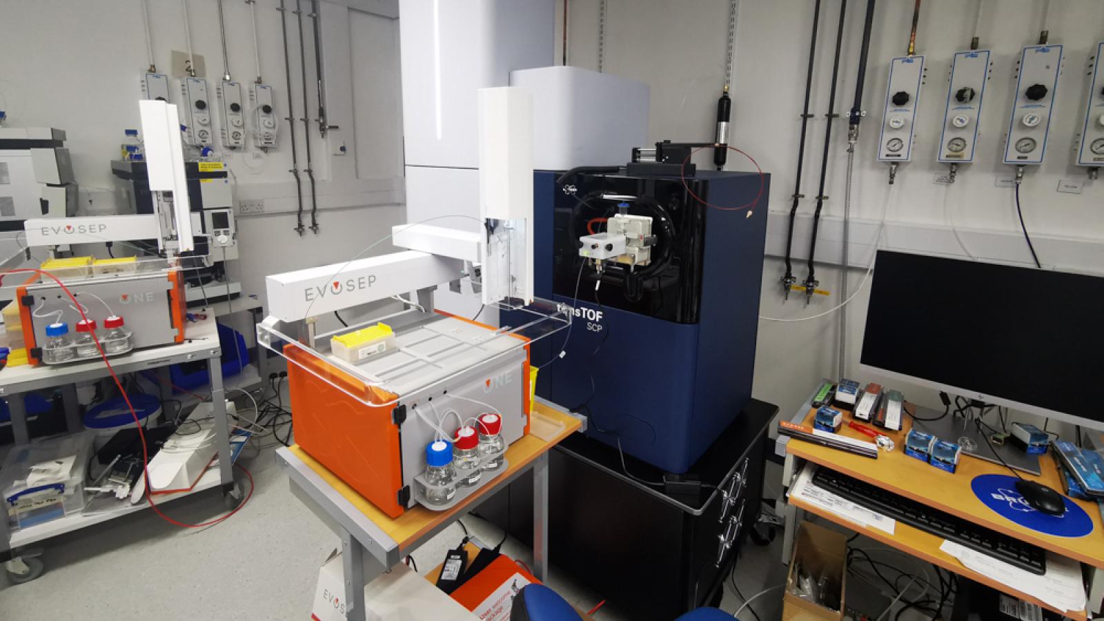A well-equipped scientific laboratory featuring various instruments and machines, including an EVOSEP system and a large blue device labeled "TimsTOF", with workstations and electrical control panels visible along the walls.